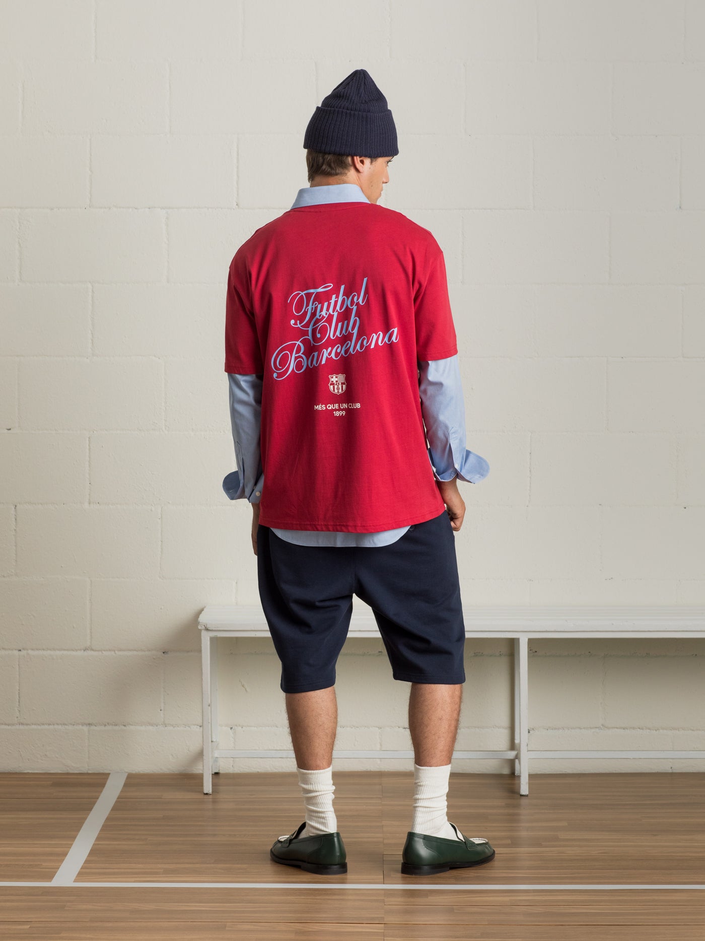 Man wearing a red 'Futbol Club Barcelona' T-shirt layered over a light blue shirt, navy shorts, and a dark beanie, facing away from the camera.