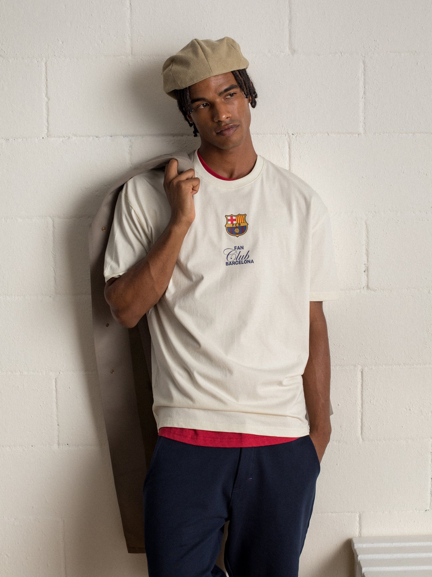 "Man wearing a beige beret and cream-colored T-shirt with FC Barcelona crest and 'Fan Club Barcelona' text, holding a light jacket over his shoulder."

