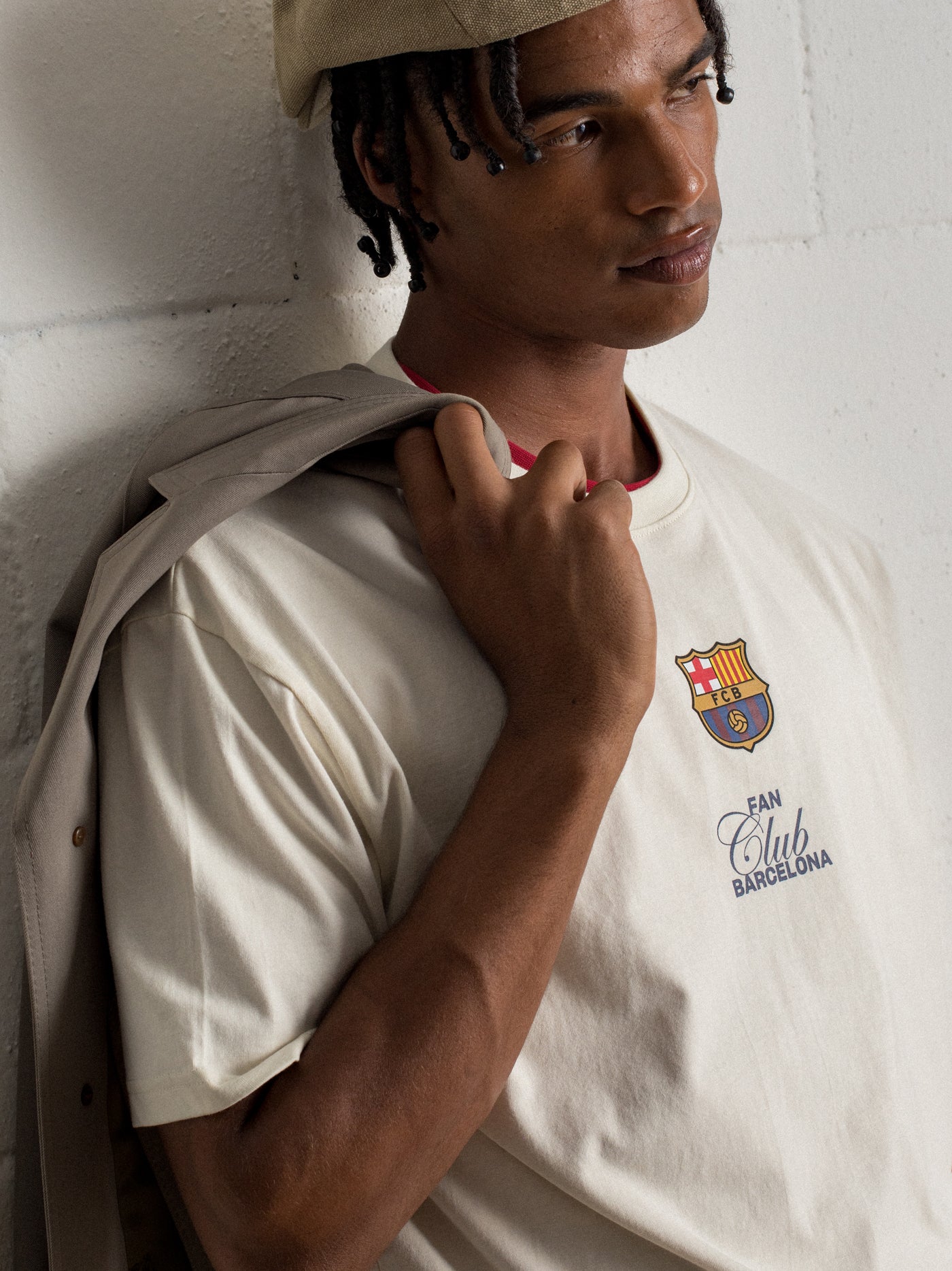 "Man wearing a beige beret and cream-colored T-shirt with FC Barcelona crest and 'Fan Club Barcelona' text, holding a light jacket over his shoulder."

