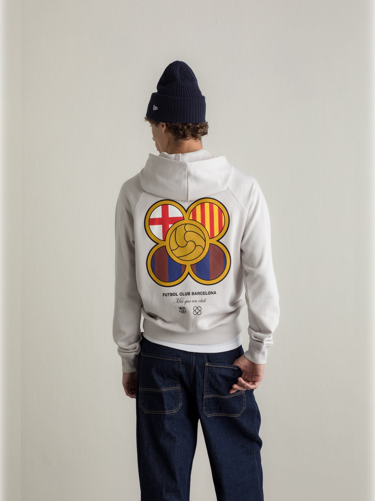 Light beige hoodie featuring a graphic of five interlocking circles with FC Barcelona colors and symbols on the back, paired with dark blue jeans.