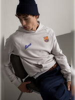 Man wearing a light beige hoodie with the Barça crest and blue 'Barça' text on the chest, paired with dark blue jeans.