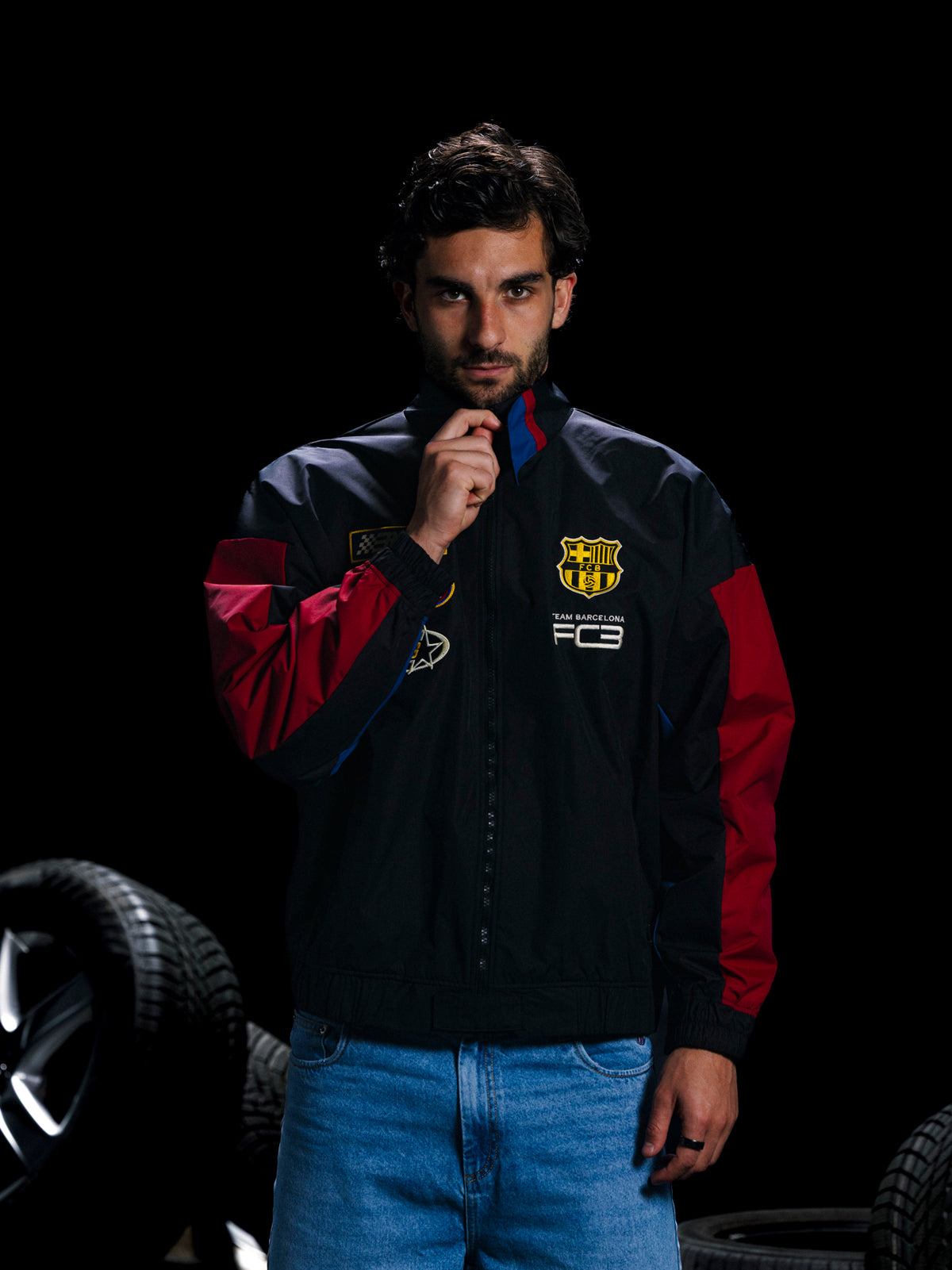 Man wearing a sports jacket with a logo, standing next to car tires on a dark background
