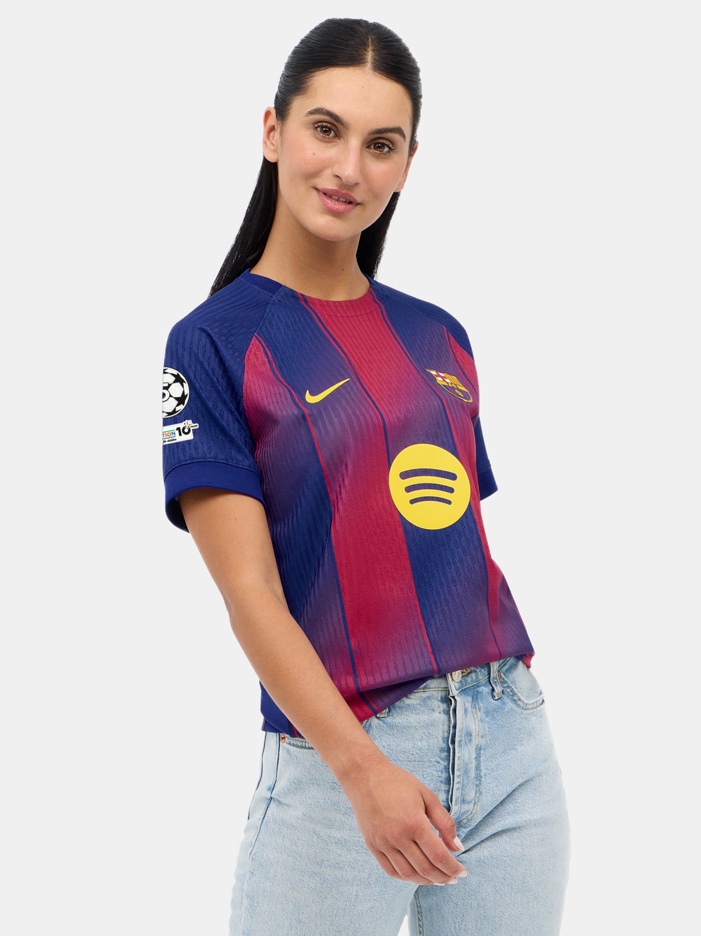 Female model smiling and slightly tilting her head while wearing the FC Barcelona 2025/26 home jersey with the Champions League badge, paired with light blue jeans, against a plain white background.
