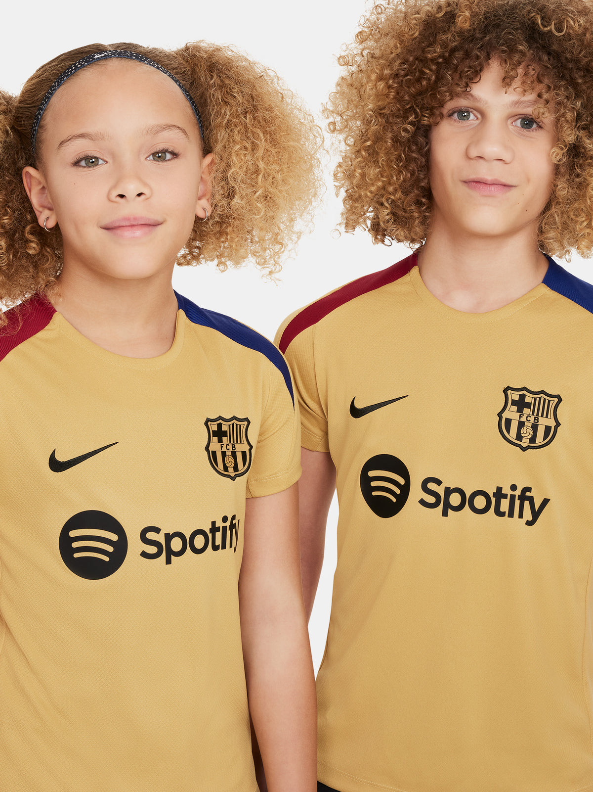 Two children wearing FC Barcelona gold training kits with short sleeves, featuring red and blue shoulder stripes, Nike logos, and Spotify sponsor branding.