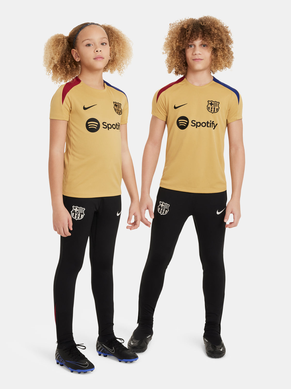 Two children wearing FC Barcelona gold training kits with short sleeves, featuring red and blue shoulder stripes, Nike logos, and Spotify sponsor branding.