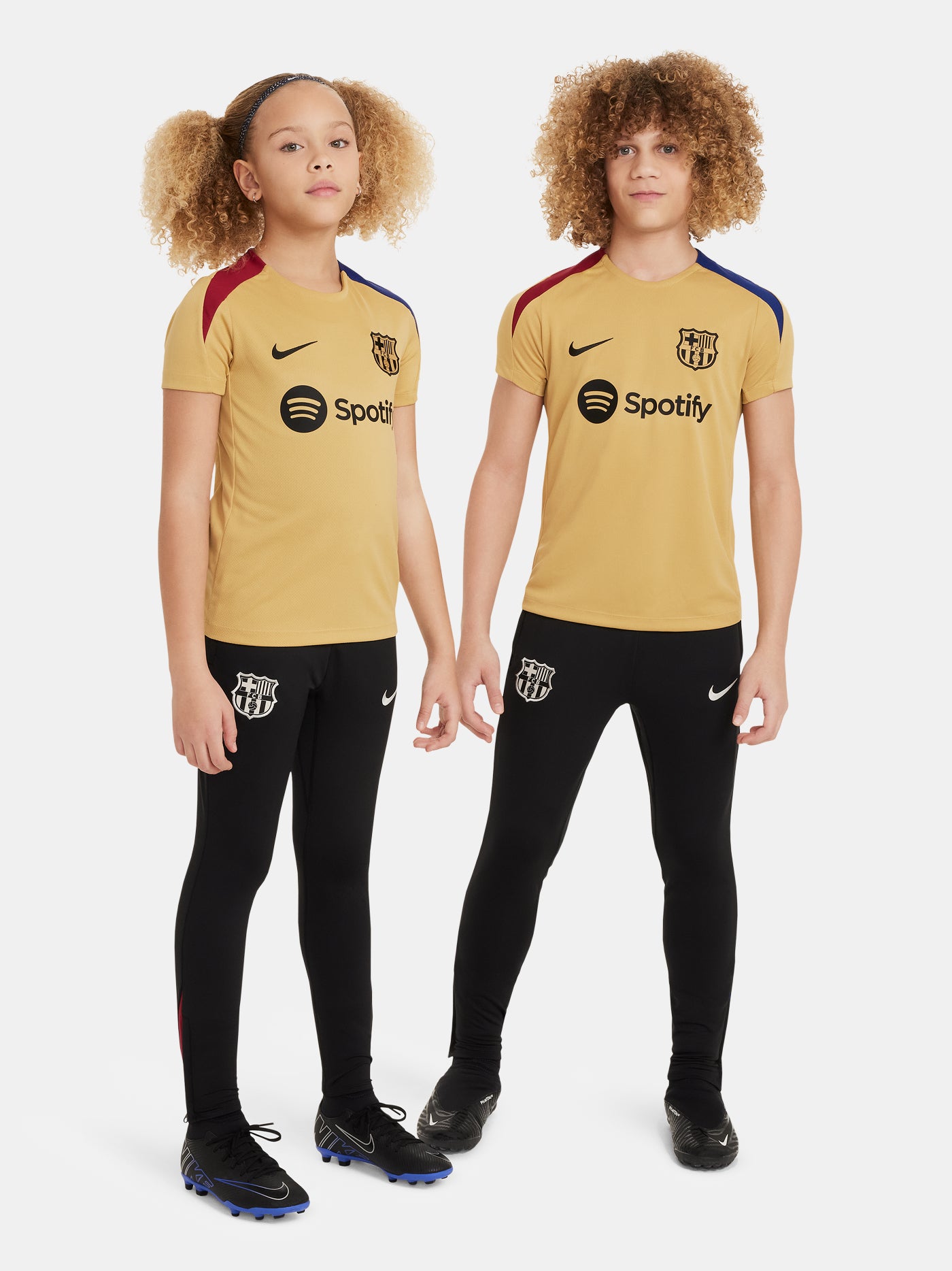 Two children wearing FC Barcelona gold training kits with short sleeves, featuring red and blue shoulder stripes, Nike logos, and Spotify sponsor branding.
