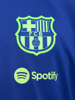FC Barcelona blue full-zip training jacket featuring neon green Nike logo on the right chest and the FC Barcelona crest with Spotify sponsor on the left chest. Black accents on the lower sleeves. Paired with black training pants.