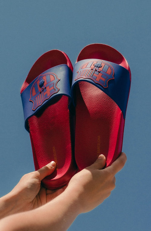 A pair of red and blue FC Barcelona slide sandals held up against a clear blue sky. The sandals feature the FC Barcelona crest prominently on the blue strap across the top.