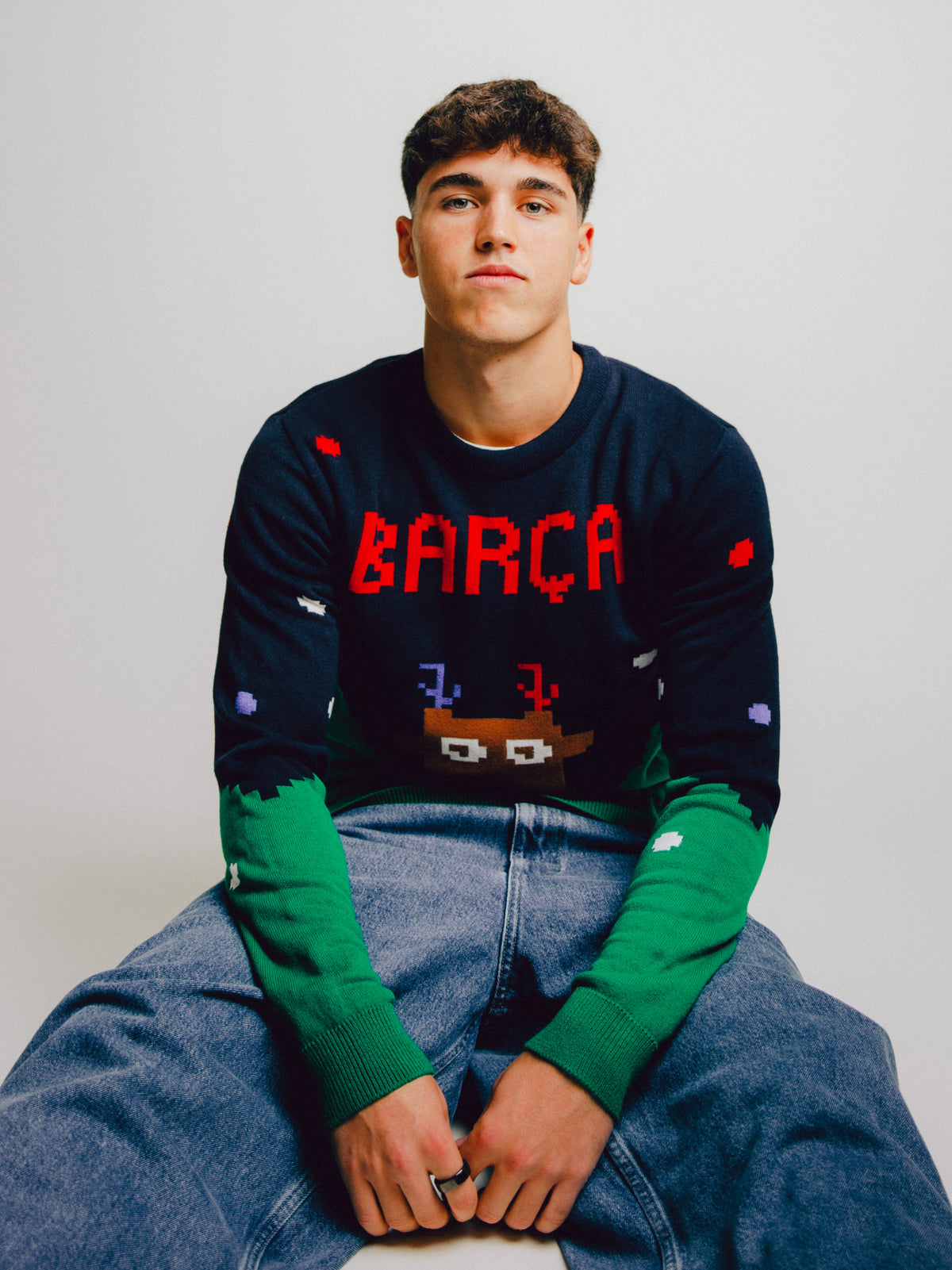 Model wearing a dark navy FC Barcelona sweater with red “BARÇA” lettering and pixel-art graphics over green sleeves and blue jeans.