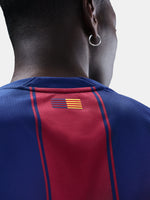 Man wearing the FC Barcelona home jersey in blue and red vertical stripes with black collar, featuring the Nike logo, club crest, and Assistència Sanitària sponsor.
