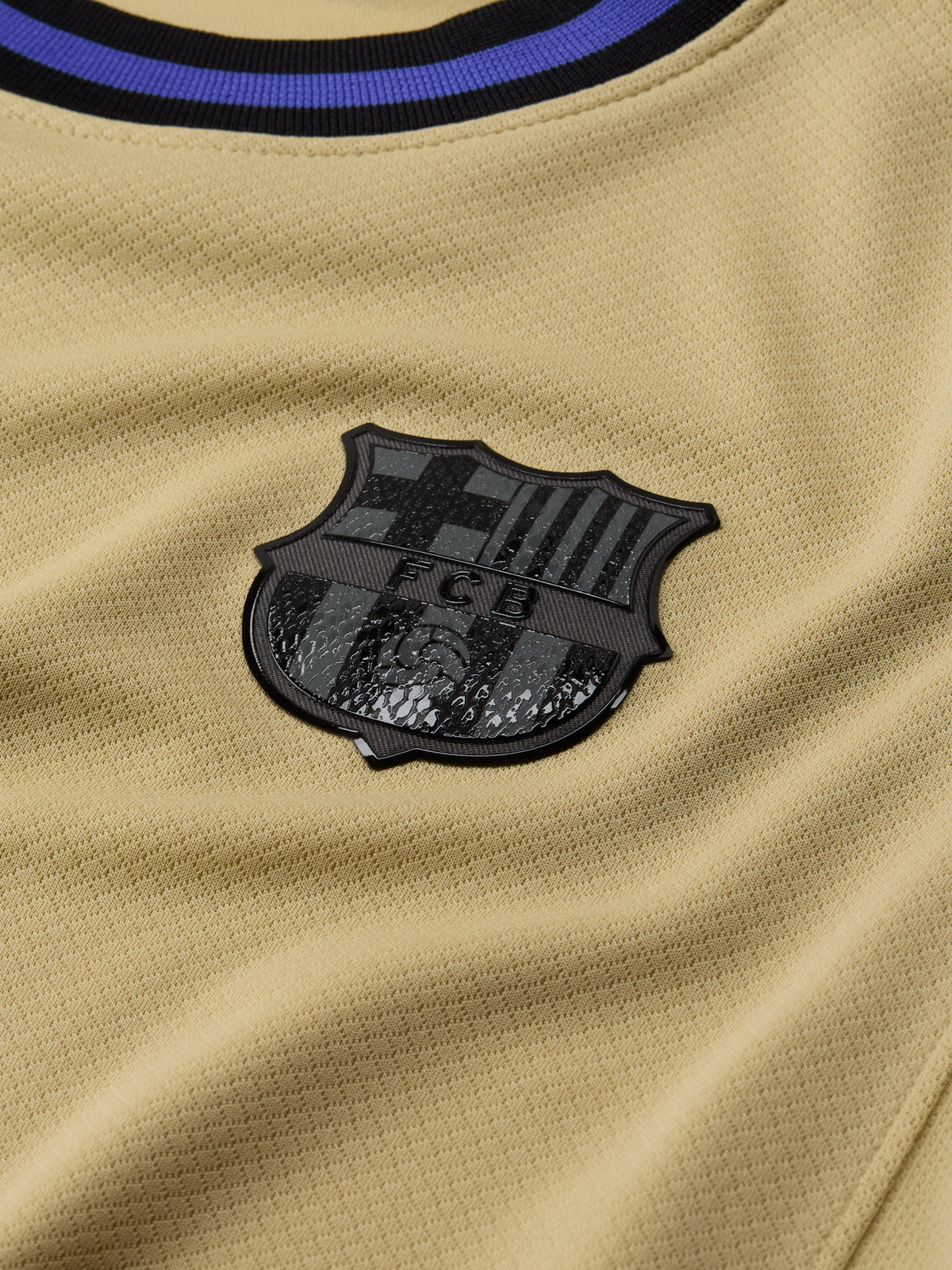 Close-up of a beige sports jersey with a black emblem featuring 'FCB' on a blurred background.