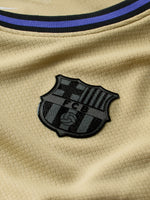 FC Barcelona kids' away kit set including beige jersey with black and purple details, black shorts with purple accents, and matching black socks, featuring club crest, Nike logo, Spotify sponsor, and Kobe Bryant tribute emblem.