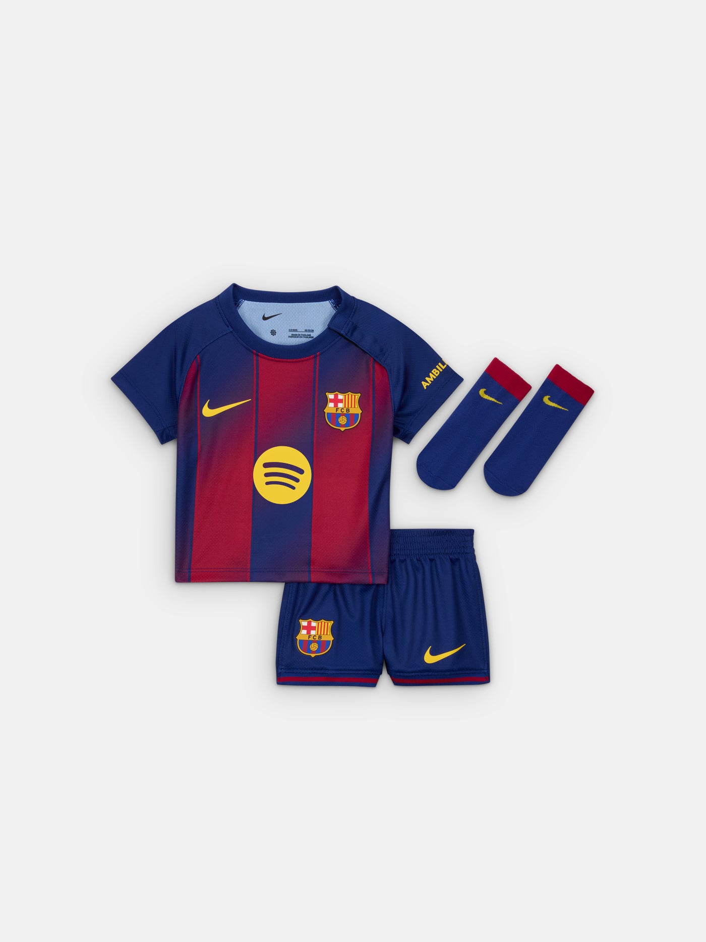 Flat lay of a Baby sized FC Barcelona home kit, including a red and blue striped jersey with Nike and Spotify logos, matching shorts with the Barça crest, and blue socks with red tops and yellow Nike swooshes.
