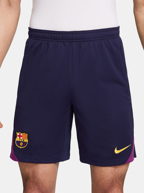 Close-up of a person wearing the FC Barcelona 2025/26 home shorts, featuring a navy blue design with the club crest on the left leg and a yellow Nike logo on the right, shown against a white background.