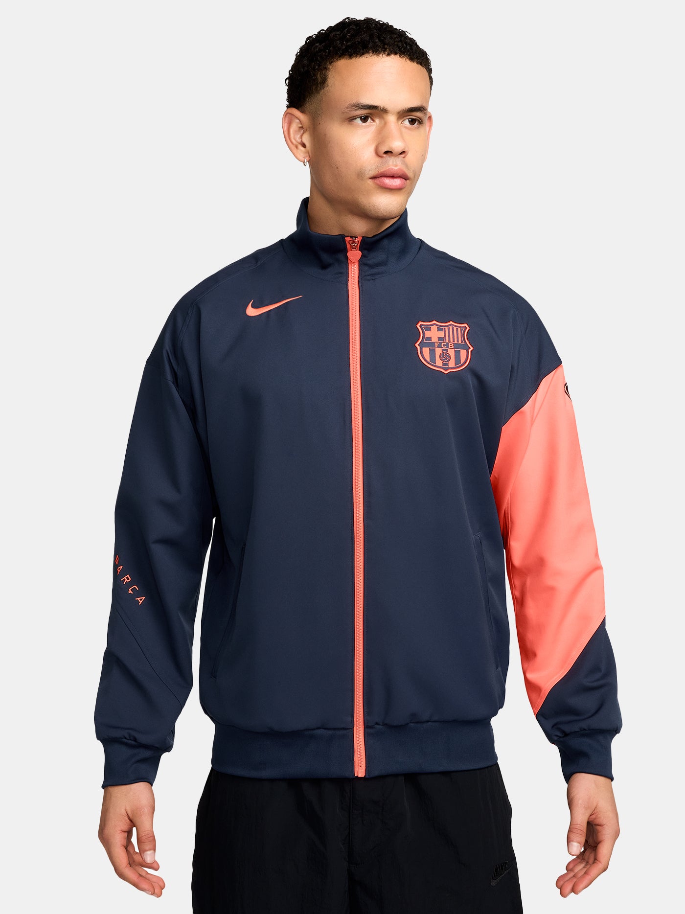 FC Barcelona navy blue jacket with coral details, including a coral sleeve, zipper, Nike logo, and club crest, paired with white Nike pants.