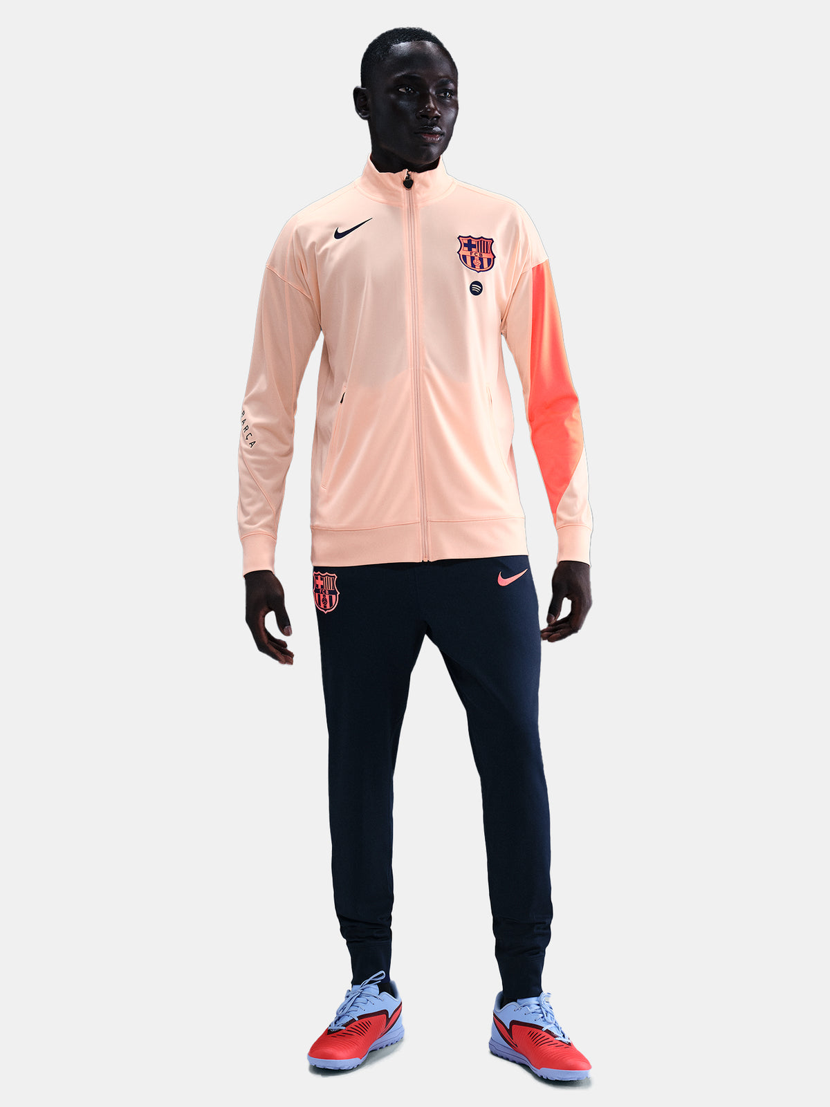 FC Barcelona Nike jacket with coral sleeve detail, paired with navy jogger pants featuring the club crest and Nike swoosh