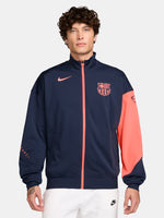FC Barcelona navy blue jacket with coral details, including a coral sleeve, zipper, Nike logo, and club crest, paired with white Nike pants.