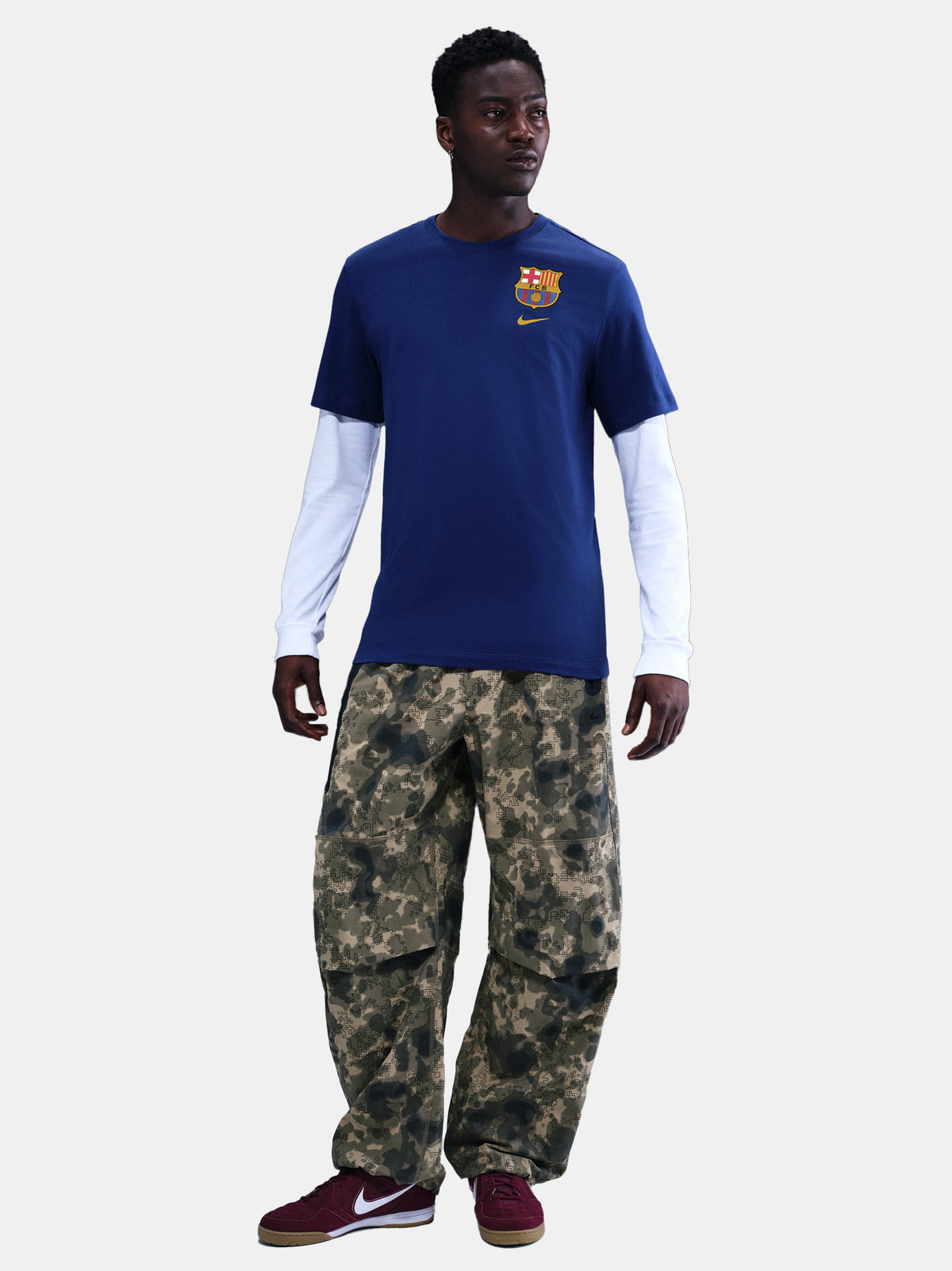 Blue Barça tee with yellow Nike logo, layered over white long sleeves and camo pants. Urban sporty style.
