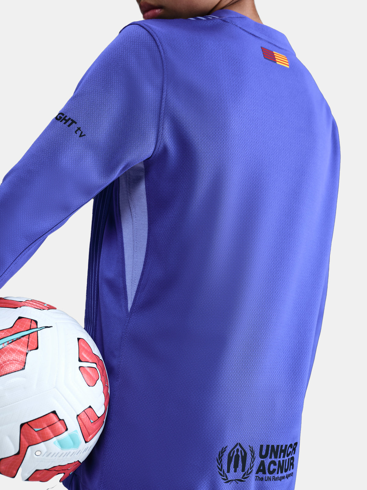 Person wearing a blue sports jersey with a logo, holding a soccer ball on a plain background