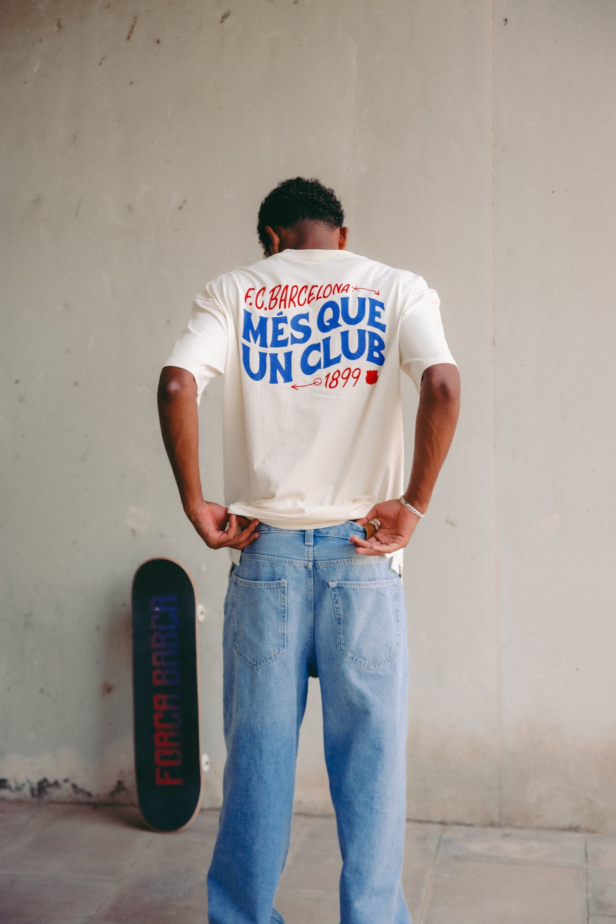 Lamine Yamal wearing an off-white FC Barcelona T-shirt with 'MÉS QUE UN CLUB' and '1899' printed on the back in red and blue, paired with light blue jeans. A skateboard with 'Força Barça' leans against the wall.