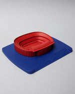 Red 3D miniature model of FC Barcelona’s Camp Nou stadium, placed on a blue surface. The model features detailed tiers, seating sections, and architectural elements of the iconic venue