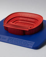 Red 3D miniature model of FC Barcelona’s Camp Nou stadium, placed on a blue surface. The model features detailed tiers, seating sections, and architectural elements of the iconic venue