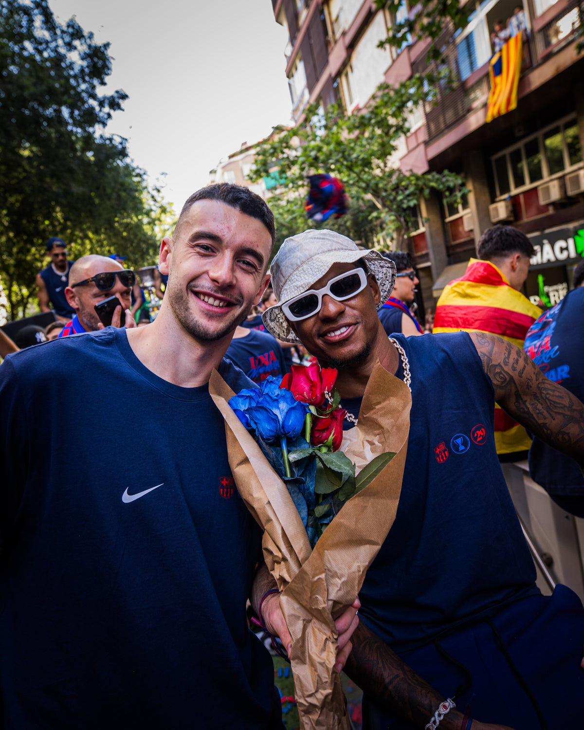 Two men standing together, one holding flowers, with a crowd and buildings in the background.
