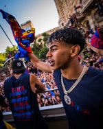Man in a blue shirt with a gold chain in a crowd, holding a colorful flag.