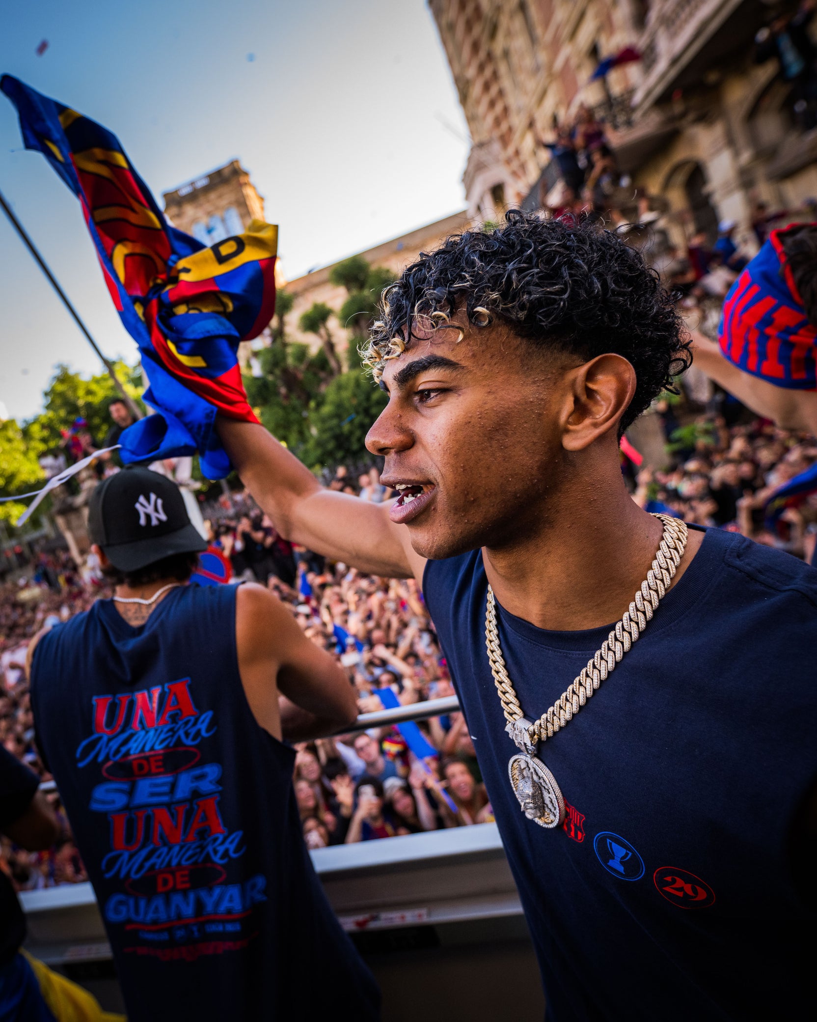 Man in a blue shirt with a gold chain in a crowd, holding a colorful flag.