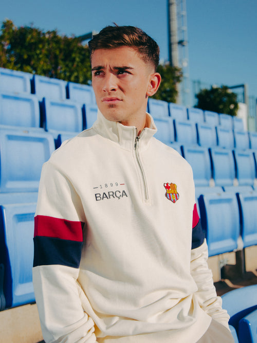 Close-up of Gavi seated in a stadium wearing a cream-colored Barça sweatshirt with red and navy sleeve stripes and embroidered club crest, evoking a calm and stylish atmosphere.
