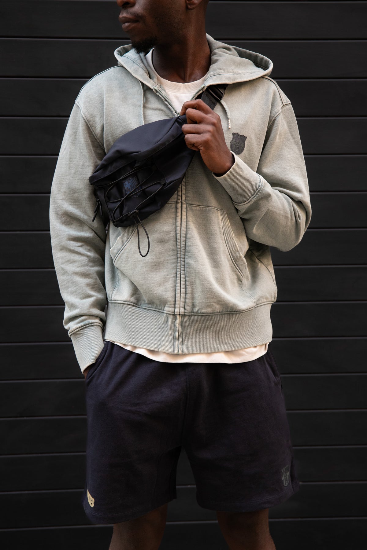 Model wearing a light grey FC Barcelona hoodie with tonal crest, paired with black shorts featuring embroidered crest, and carrying a black crossbody bag