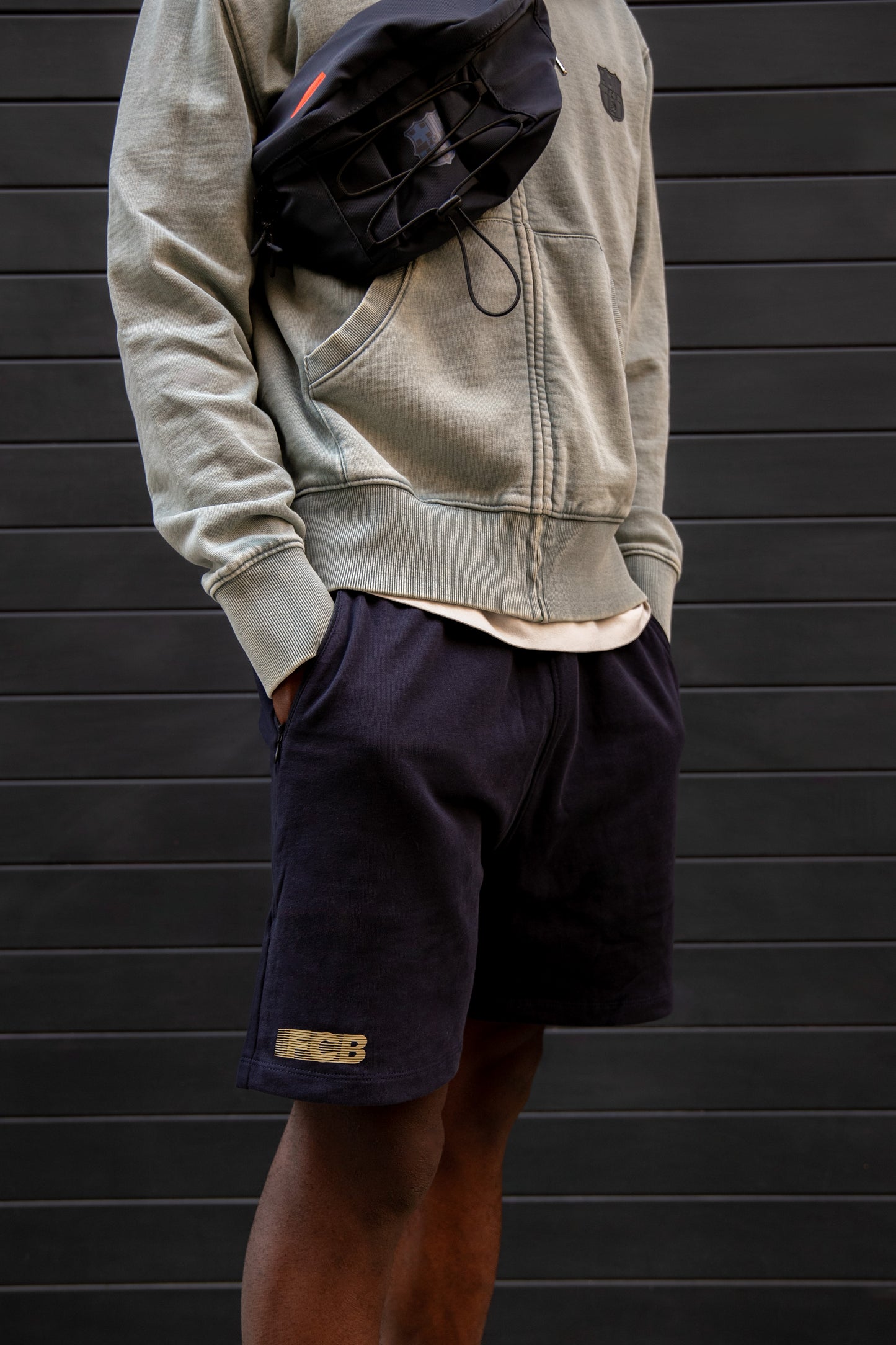 Model wearing a light grey FC Barcelona hoodie with tonal crest, paired with black shorts featuring embroidered crest, and carrying a black crossbody bag