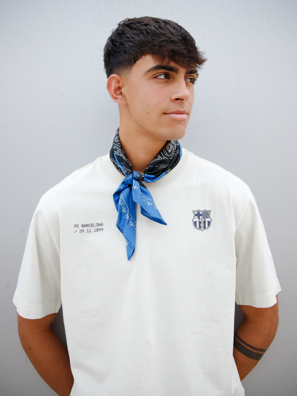 Marc Bernal wearing an off-white FC Barcelona T-shirt with tonal club crest and date detail on the chest, paired with a blue and black patterned neck scarf