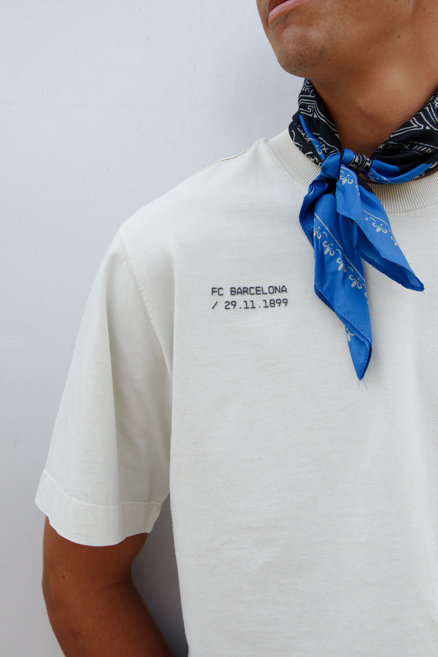 Marc Bernal wearing an off-white FC Barcelona T-shirt with tonal club crest and date detail on the chest, paired with a blue and black patterned neck scarf
