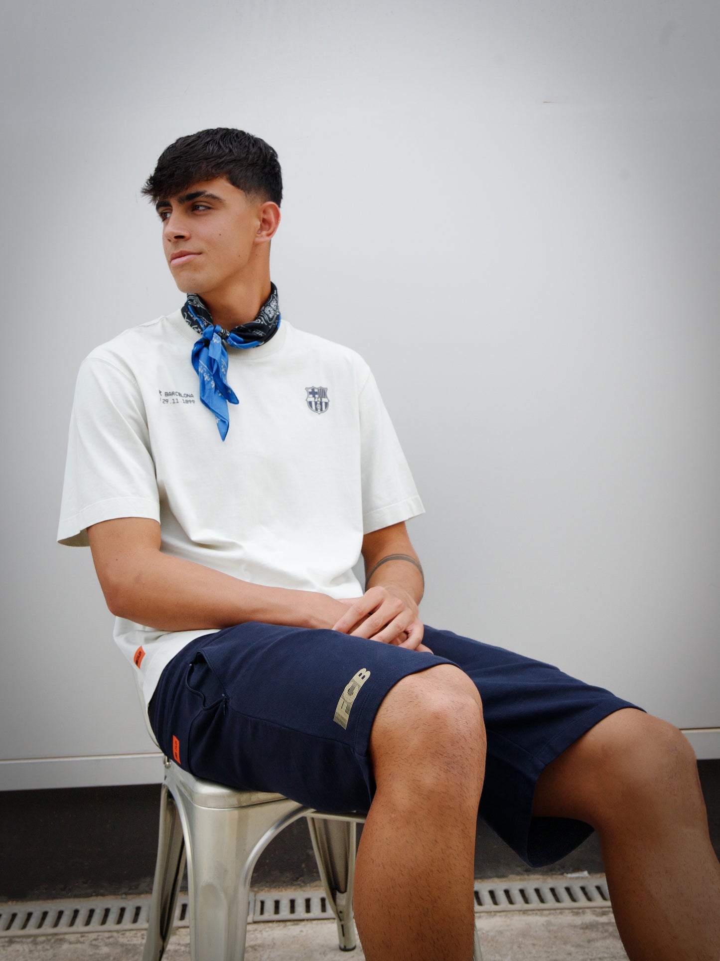Marc Bernal wearing an off-white FC Barcelona T-shirt with tonal club crest and date detail on the chest, paired with a blue and black patterned neck scarf