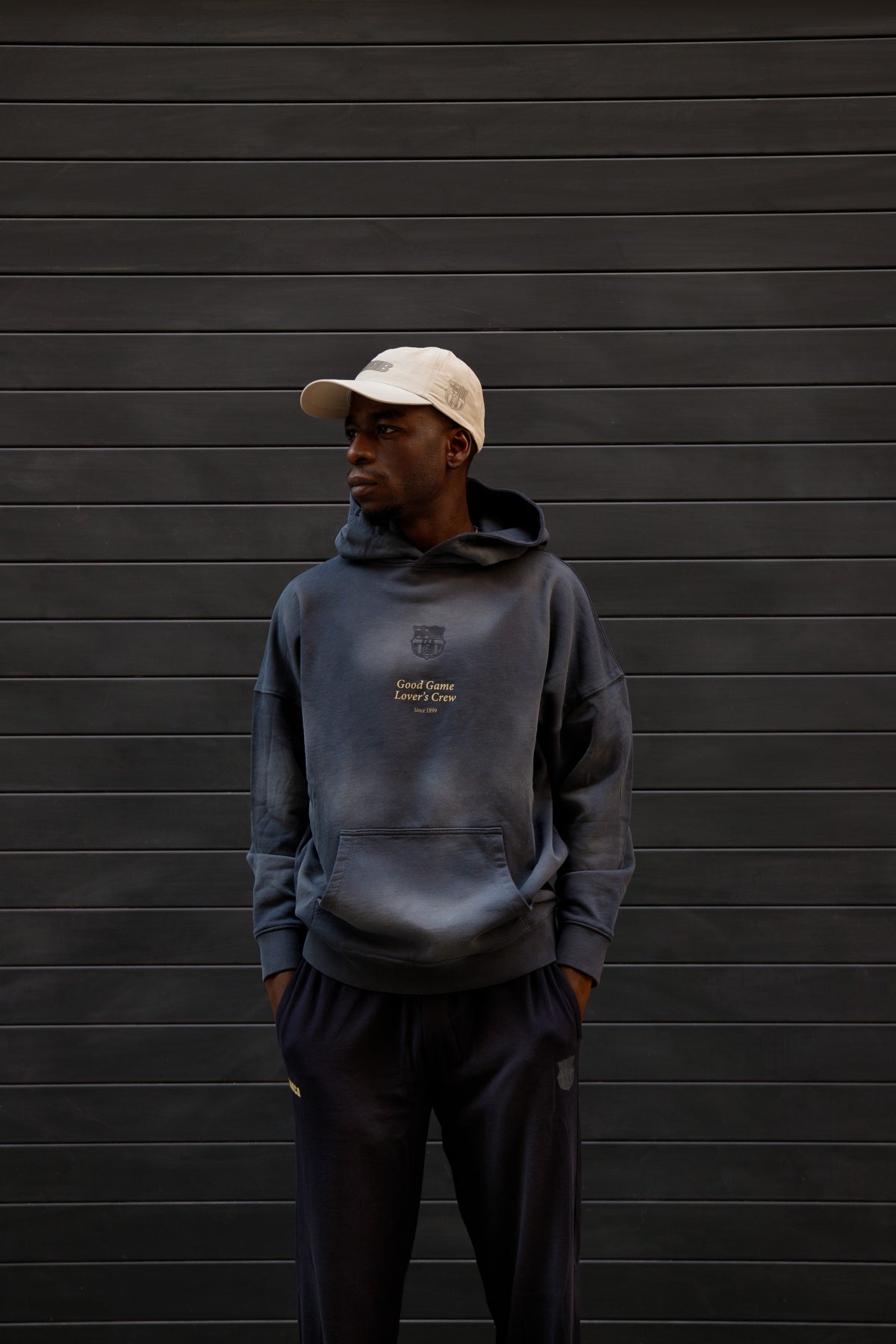 Man wearing a dark blue FC Barcelona hoodie with subtle crest and “Good Game Lover’s Crew” text on the chest, paired with matching joggers and a beige cap, standing against a dark background.