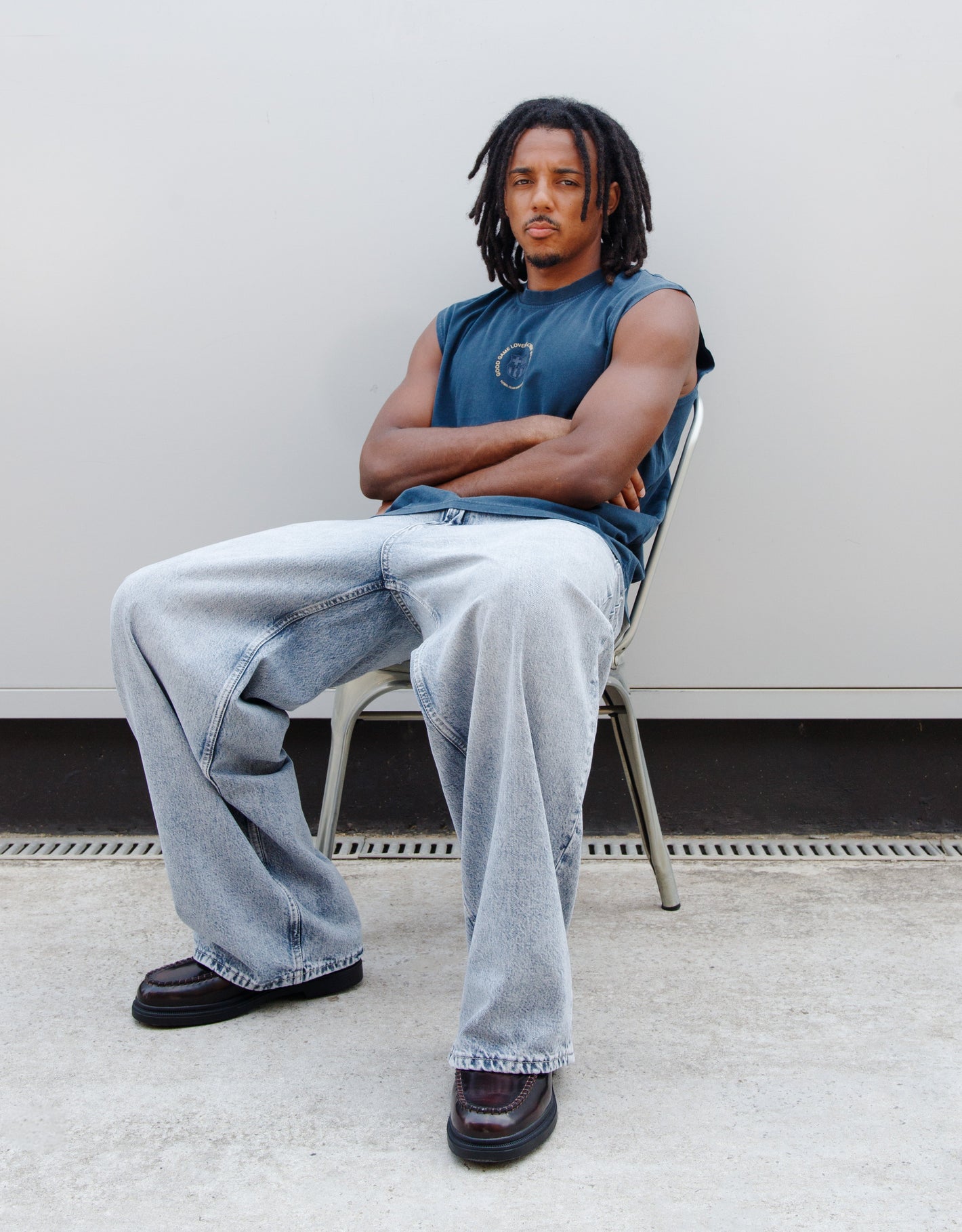 Man sitting on a chair wearing a blue sleeveless shirt and light blue jeans against a plain background