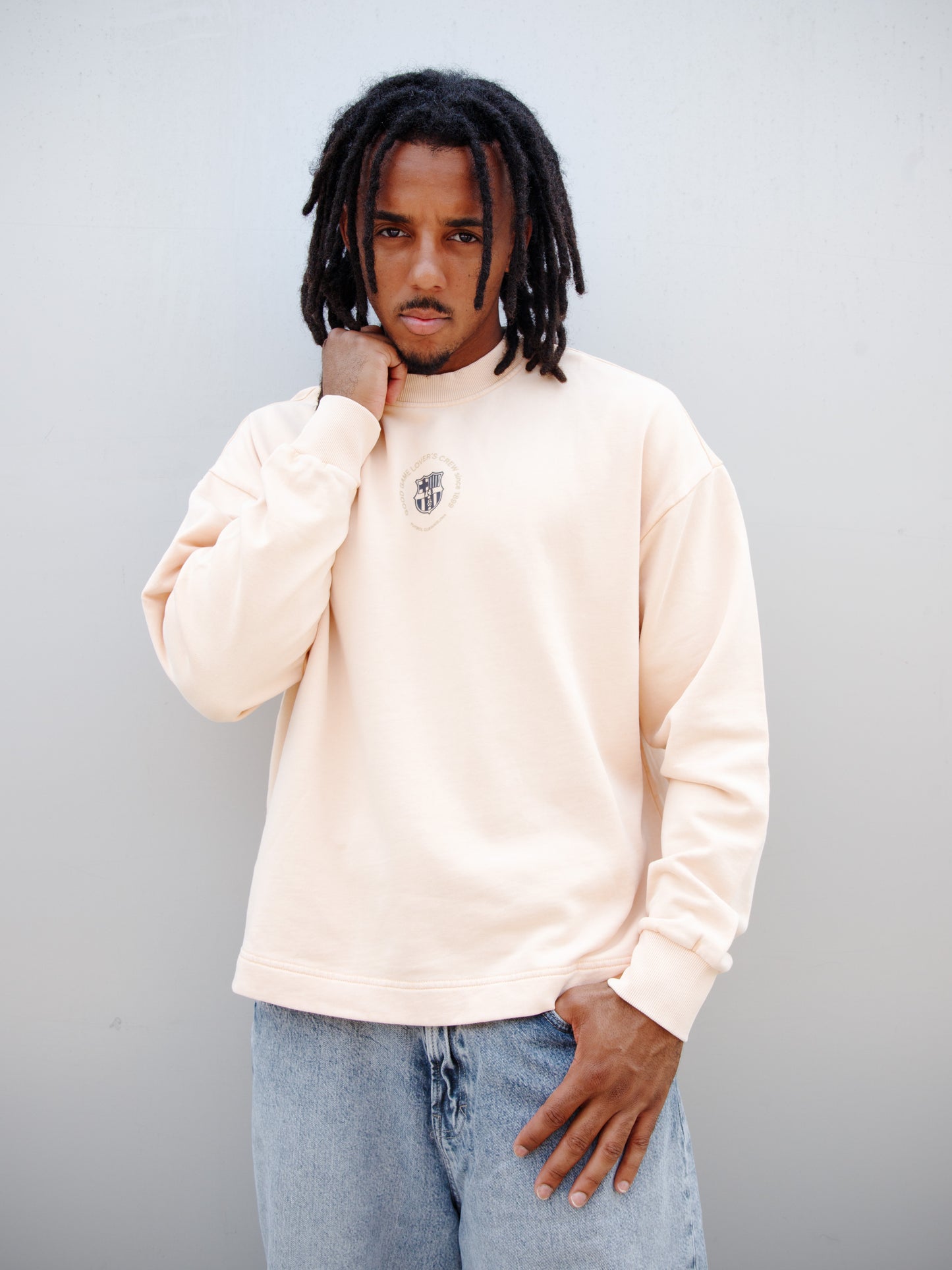 Jules Kounde wearing a light peach FC Barcelona sweatshirt with circular crest print on the chest, paired with light blue denim jeans.