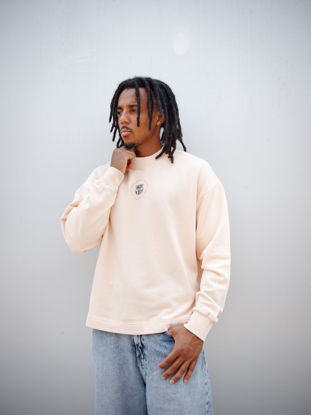 Jules Kounde wearing a light peach FC Barcelona sweatshirt with circular crest print on the chest, paired with light blue denim jeans.