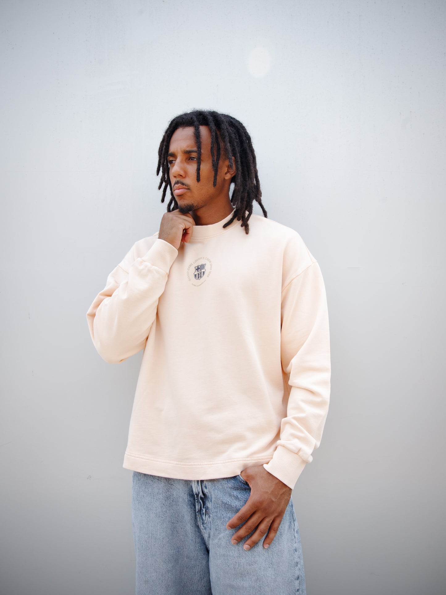 Jules Kounde wearing a light peach FC Barcelona sweatshirt with circular crest print on the chest, paired with light blue denim jeans.