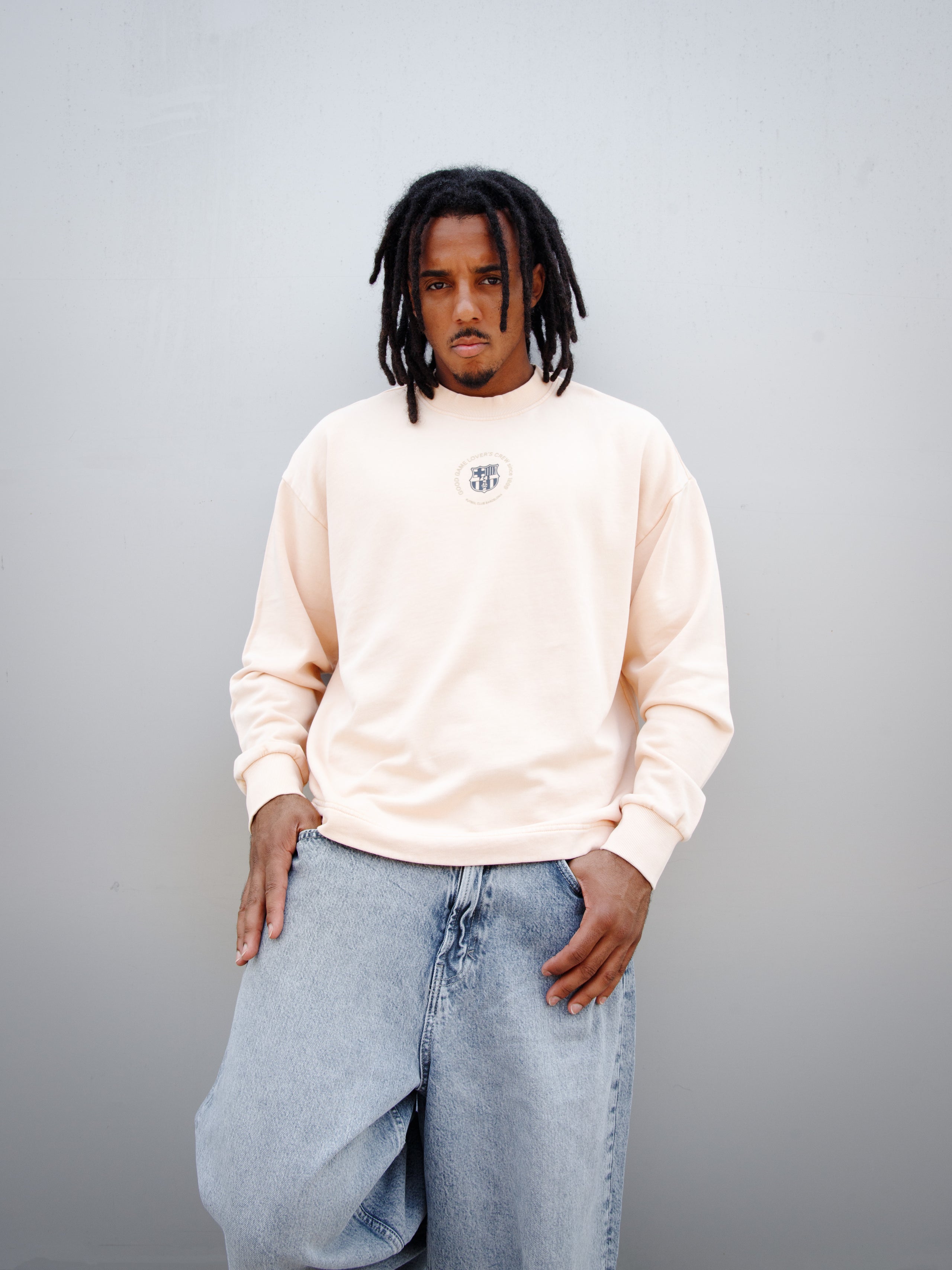 Jules Kounde wearing a light peach FC Barcelona sweatshirt with circular crest print on the chest, paired with light blue denim jeans.
