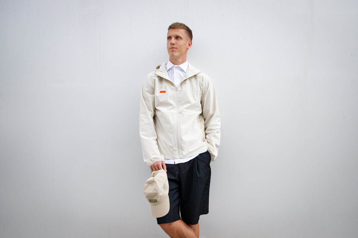 Dani Olmo wearing a light beige FC Barcelona hooded jacket with subtle crest and orange detail, paired with black shorts, white polo, and holding a matching beige cap