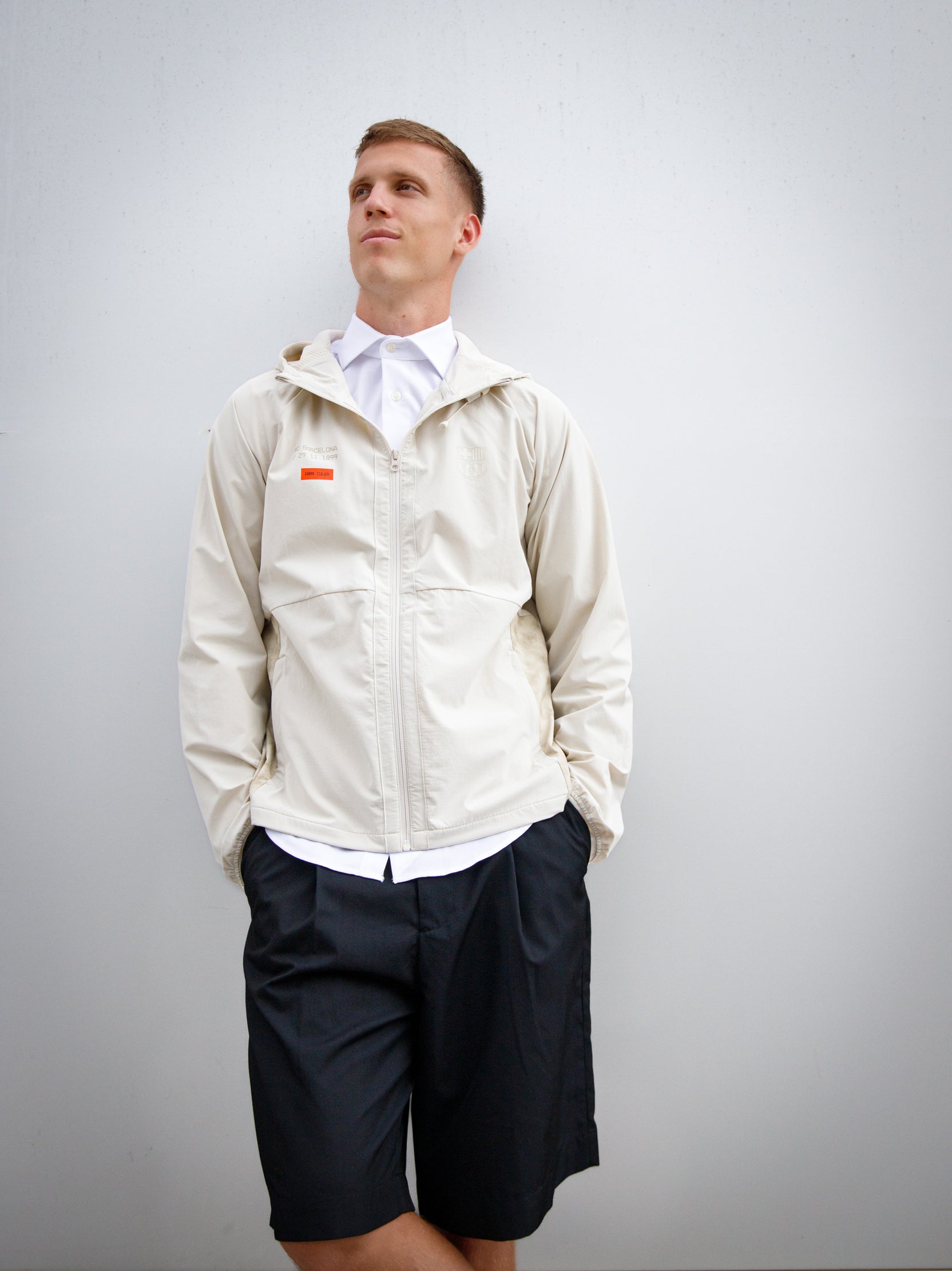 Dani Olmo wearing a light beige FC Barcelona hooded jacket with subtle crest and orange detail, paired with black shorts and white polo shirt