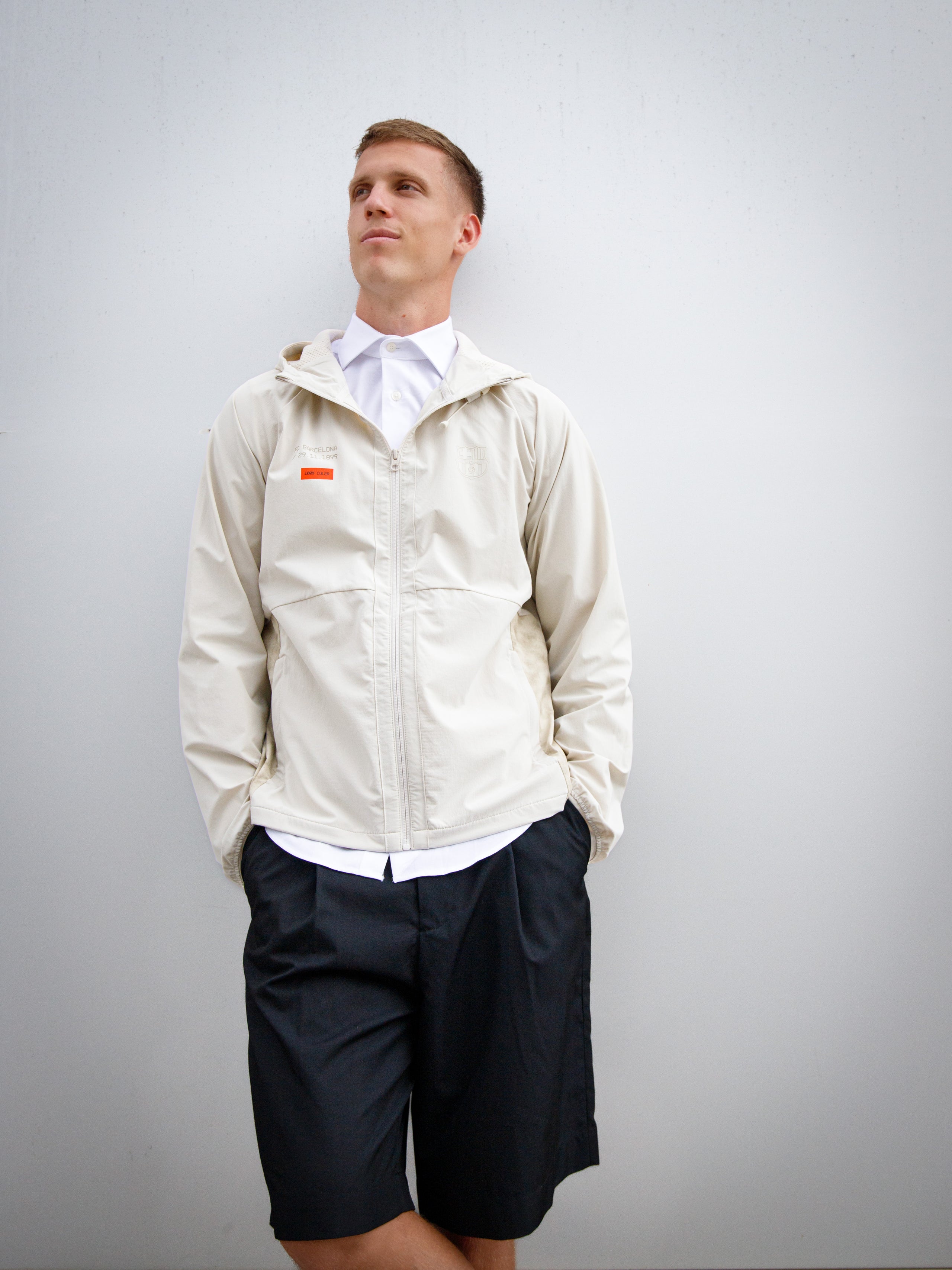 Dani Olmo wearing a light beige FC Barcelona hooded jacket with subtle crest and orange detail, paired with black shorts and white polo shirt