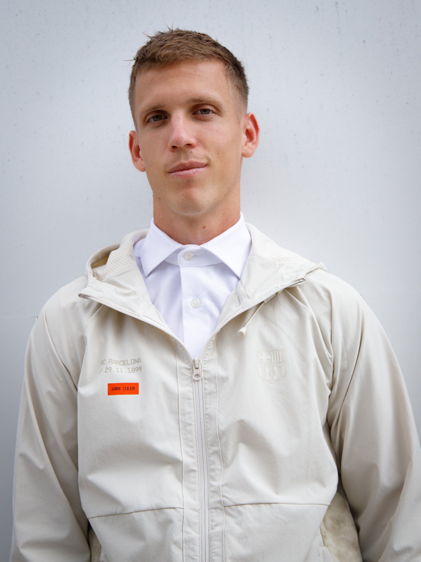 Dani Olmo wearing a light beige FC Barcelona hooded jacket with subtle crest and orange detail, paired with black shorts and white polo shirt
