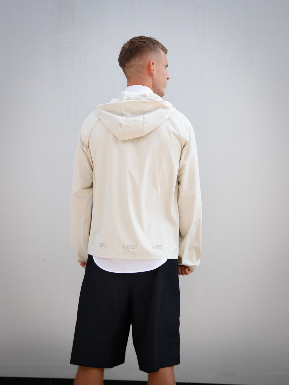 Dani Olmo wearing a light beige FC Barcelona hooded jacket with subtle crest and orange detail, paired with black shorts and white polo shirt