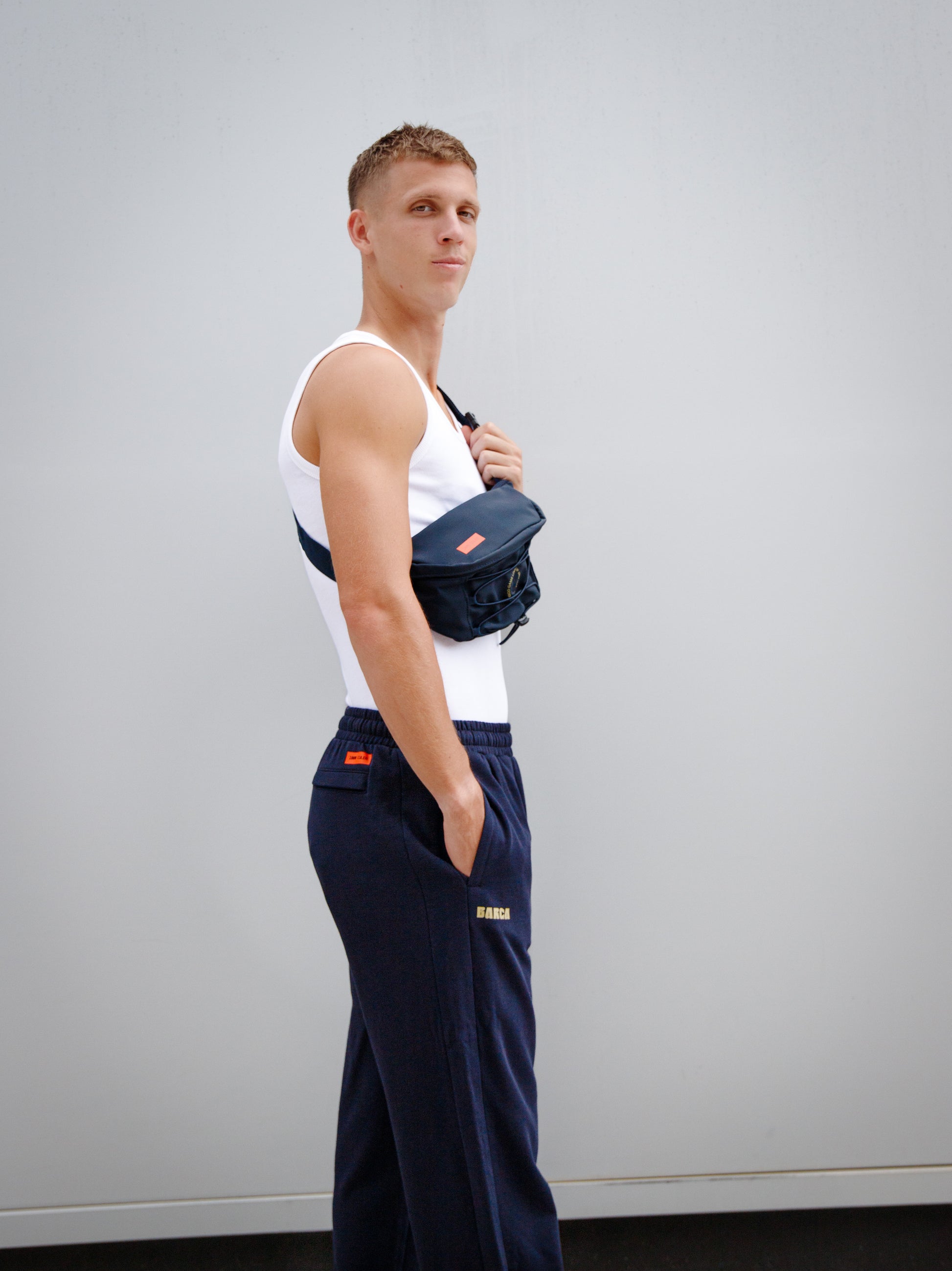 Model wearing a white tank top and navy blue FC Barcelona sweatpants with golden 'Barça' text, paired with a navy waist bag featuring crest and orange label