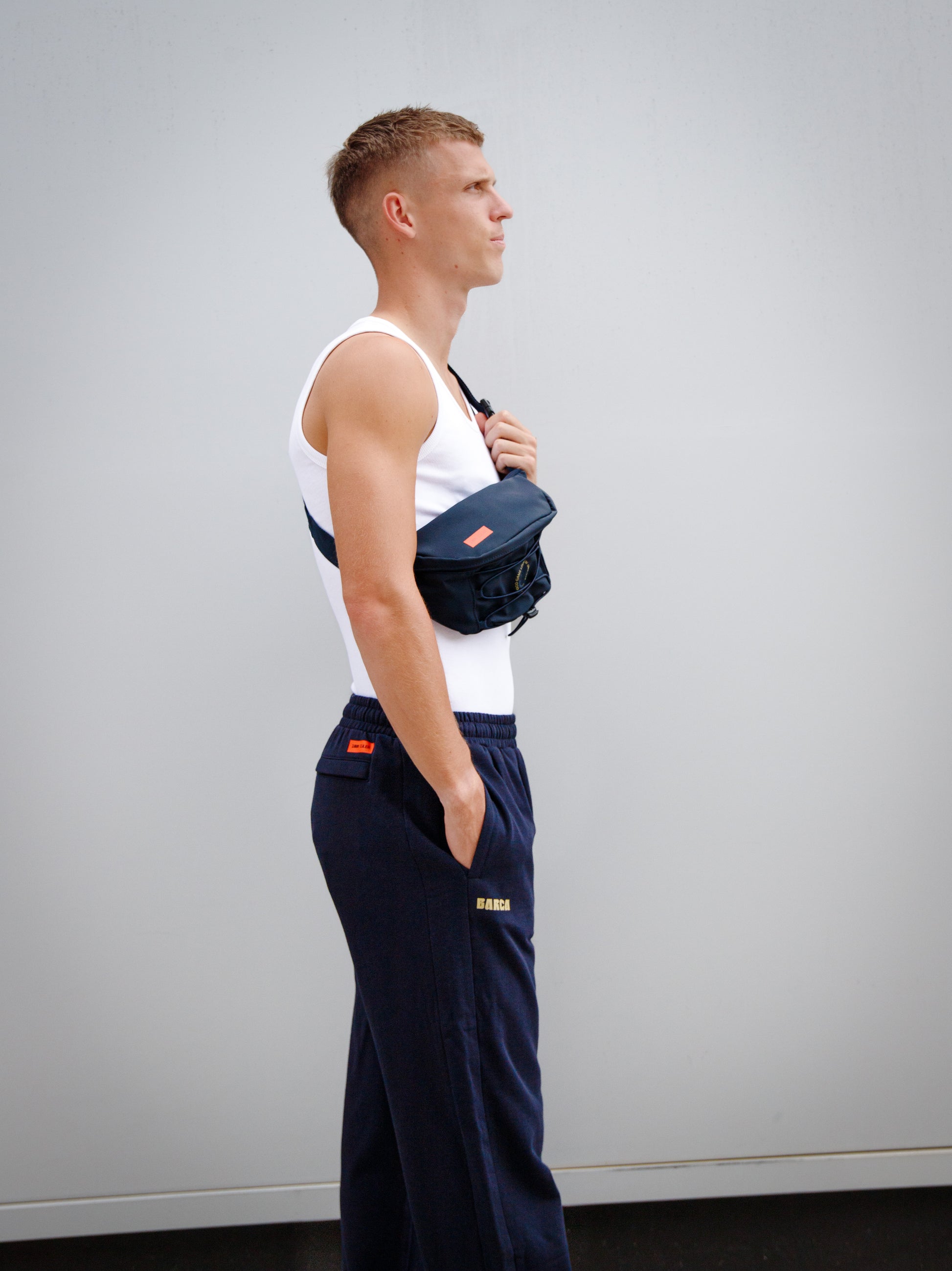 Model wearing a white tank top and navy blue FC Barcelona sweatpants with golden 'Barça' text, paired with a navy waist bag featuring crest and orange label