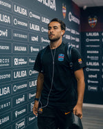 Modèle portant une tenue d’entraînement noire FC Barcelona avec l’écusson du club et le logo Nike, marchant dans un couloir brandé LaLiga avec des écouteurs et un sac à dos.