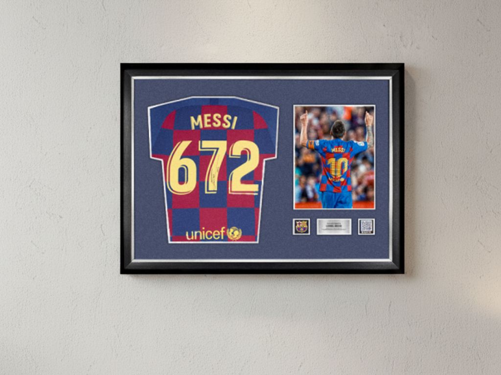 Framed FC Barcelona memorabilia featuring a Messi jersey with number 672, alongside a photo of the player celebrating, set against a blue background with club crest details.